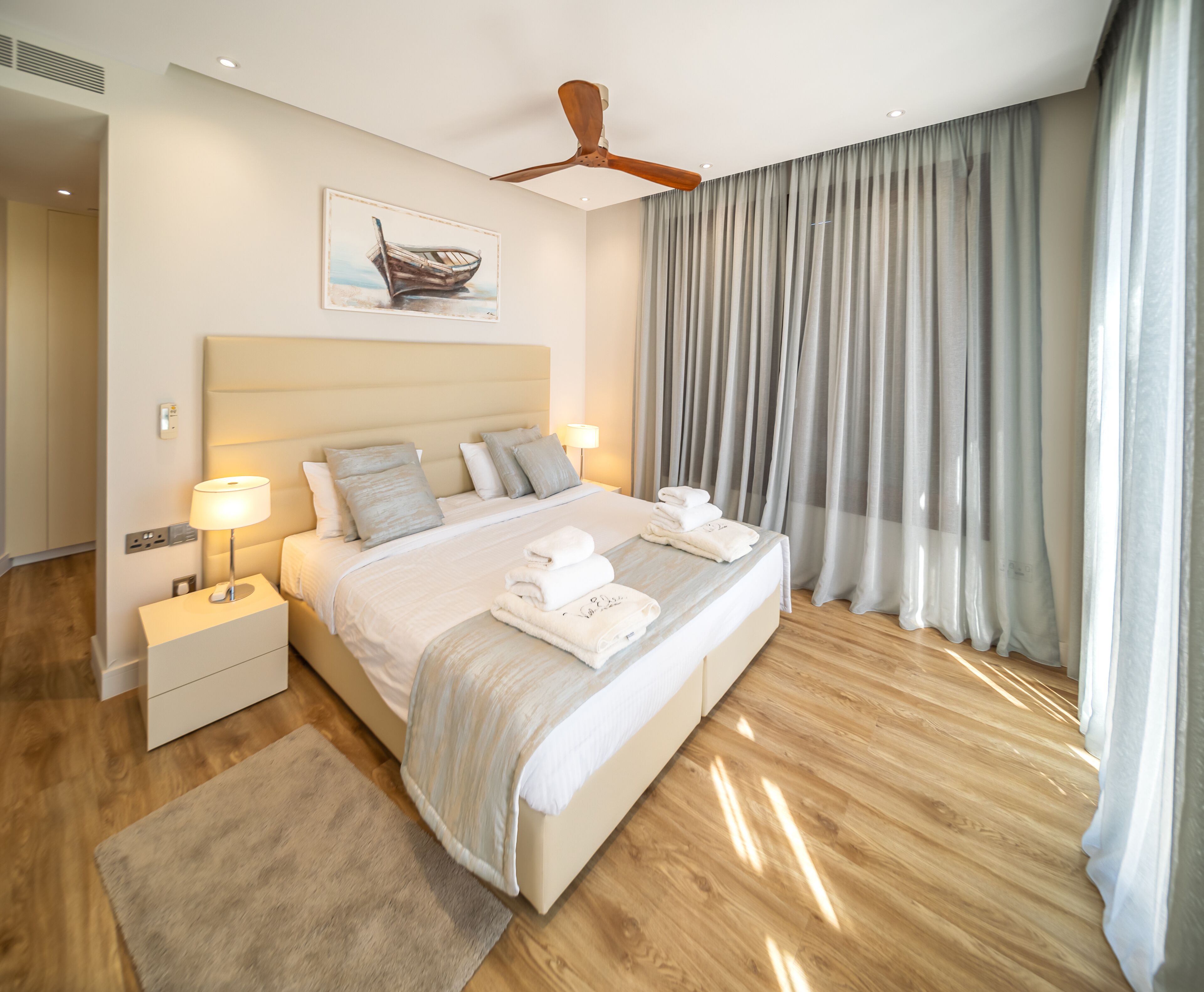 A modern hotel room with a large bed, a ceiling fan, a painting of a boat, and a view of the sea through sheer curtains.