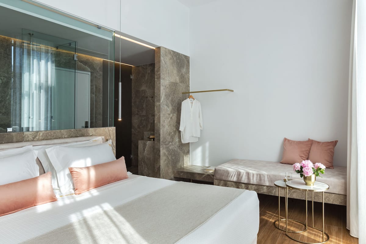 A modern hotel room with a large bed, a seating area with a bench, a small table with a vase of flowers, and a large mirror.