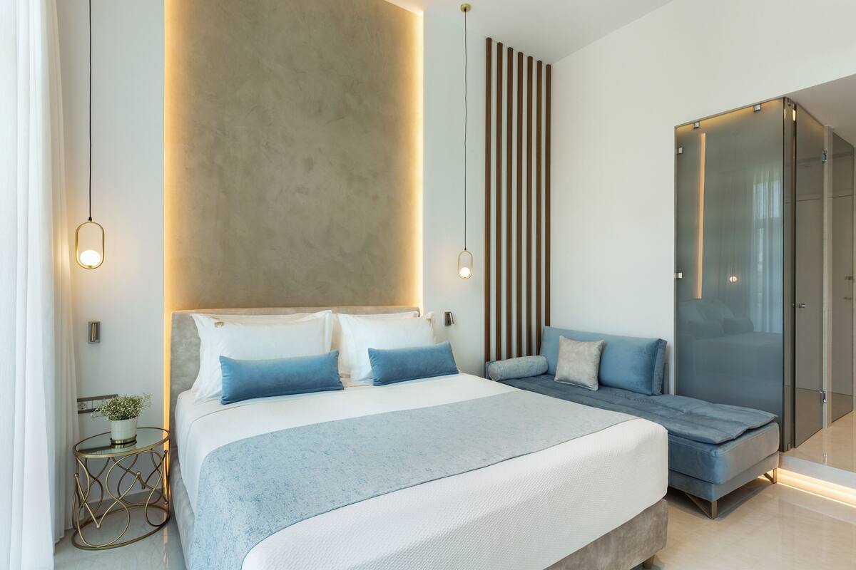 A modern bedroom with a large bed, a seating area, and a glass-enclosed walk-in closet.