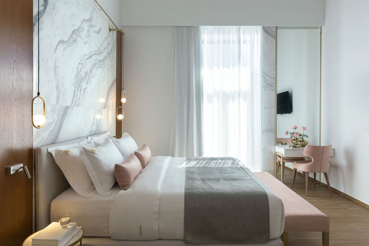 A modern hotel room with a large bed, bedside table, and a desk with a chair.