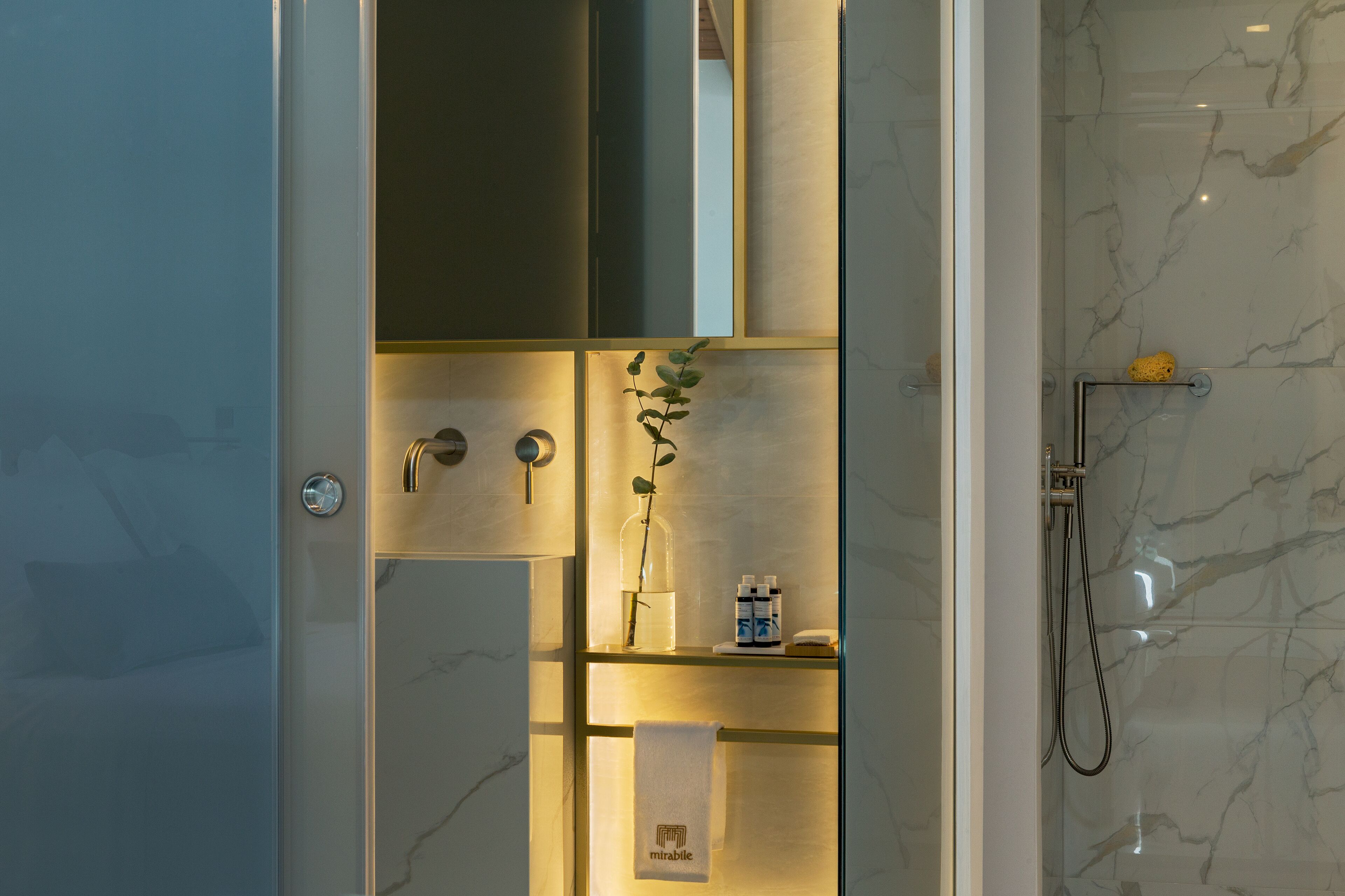 A modern bathroom with a glass shower enclosure, marble walls, and a shelf with toiletries.