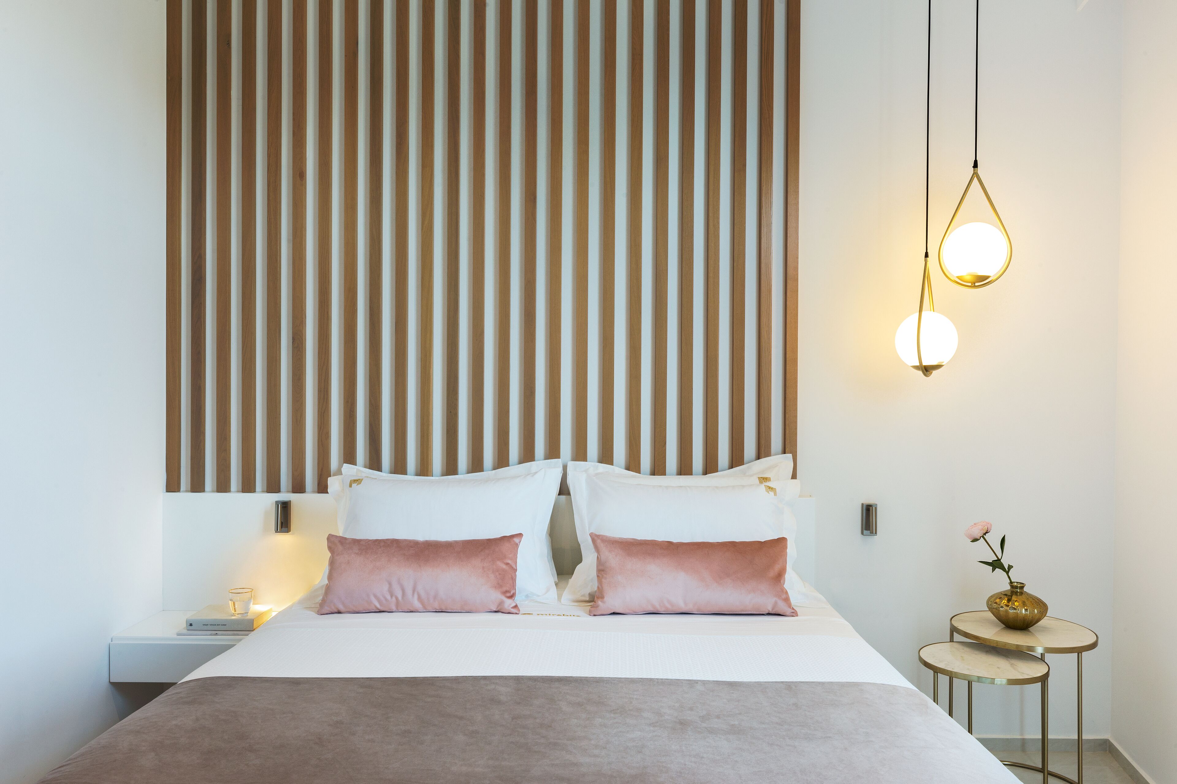 A neatly made bed with two pillows, a bedside table with a book and a glass, and a small round table with a vase and a flower.