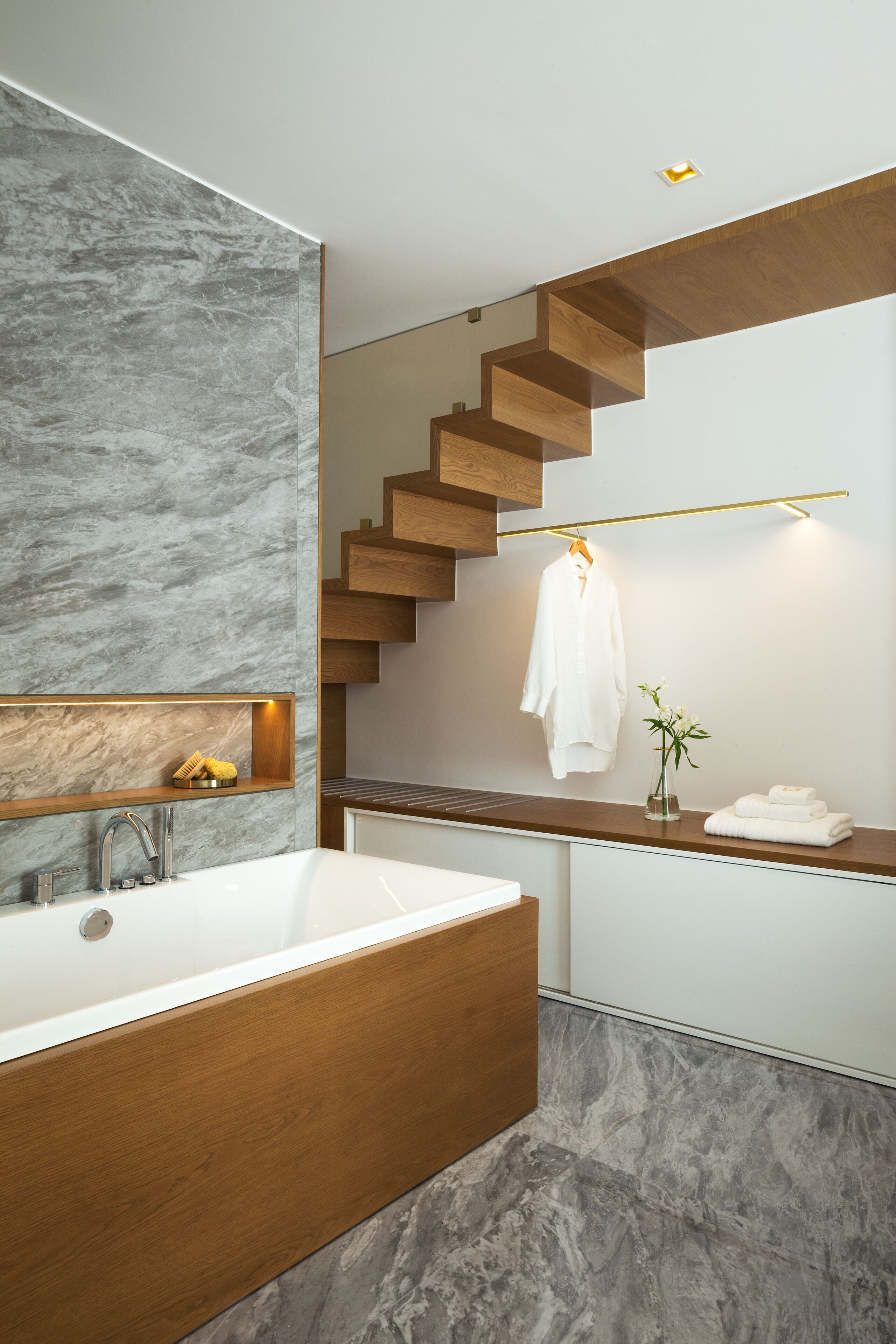 A modern bathroom with a marble wall, wooden vanity, and a built-in bathtub.