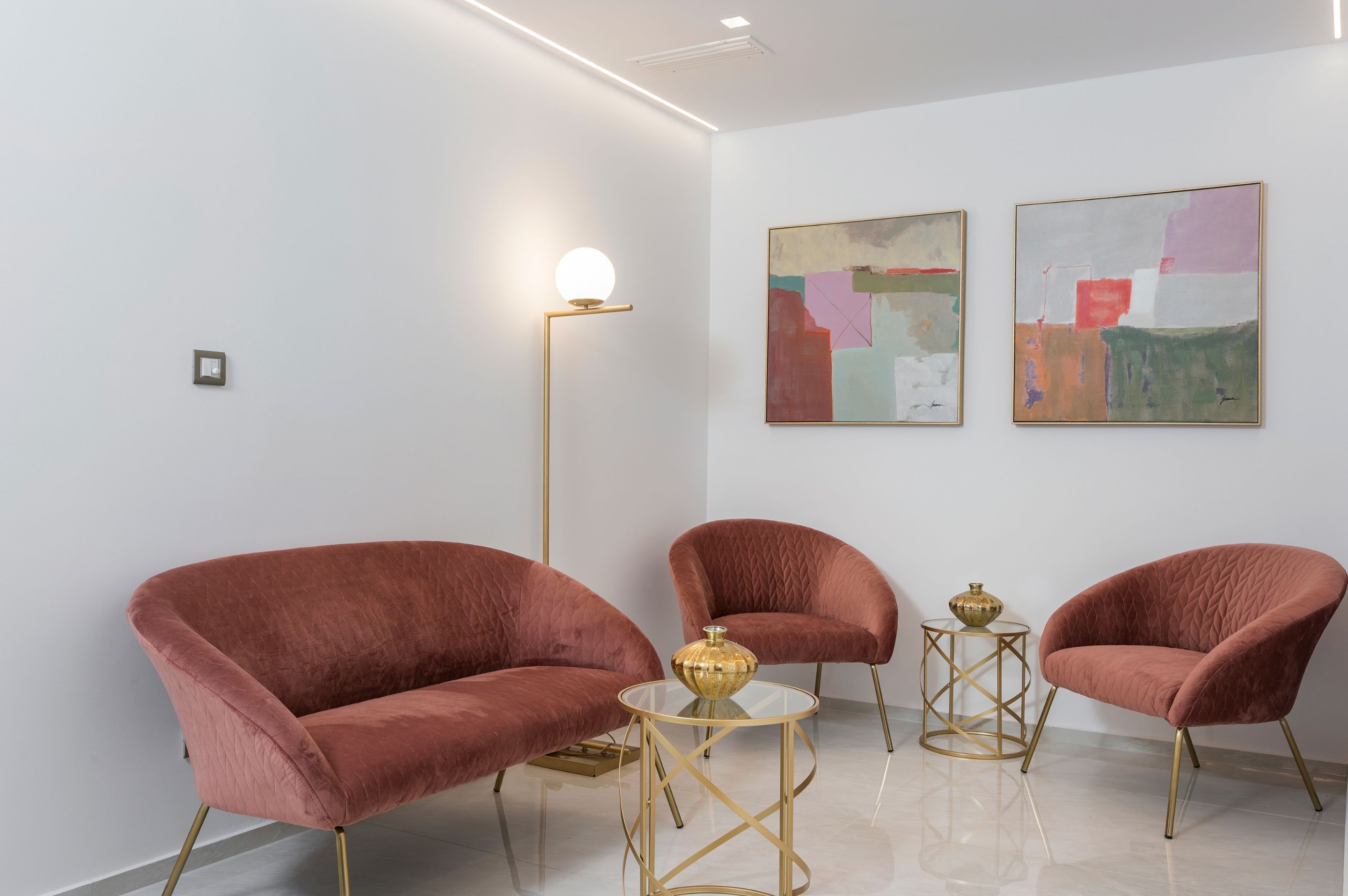 A modern room with two plush red armchairs, a small round table with a gold vase, and two abstract paintings on the wall.