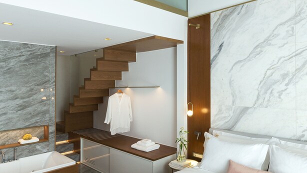 A modern hotel room with a large bed, a bathroom with a bathtub, a staircase to an upper level, and a marble wall.