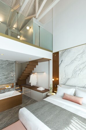 A modern hotel room with a large bed, a bathroom with a bathtub, a staircase to an upper level, and a marble wall. - Mirabile Luxury Suites (Chania)