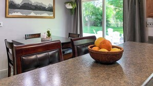 Un coin repas avec une table en granit, des chaises en cuir, un panier de fruits, un tableau représentant un paysage, et une vue sur un espace extérieur verdoyant.