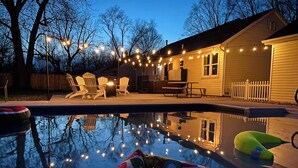 Un patio trasero con piscina, sillas de deck y luces colgantes.