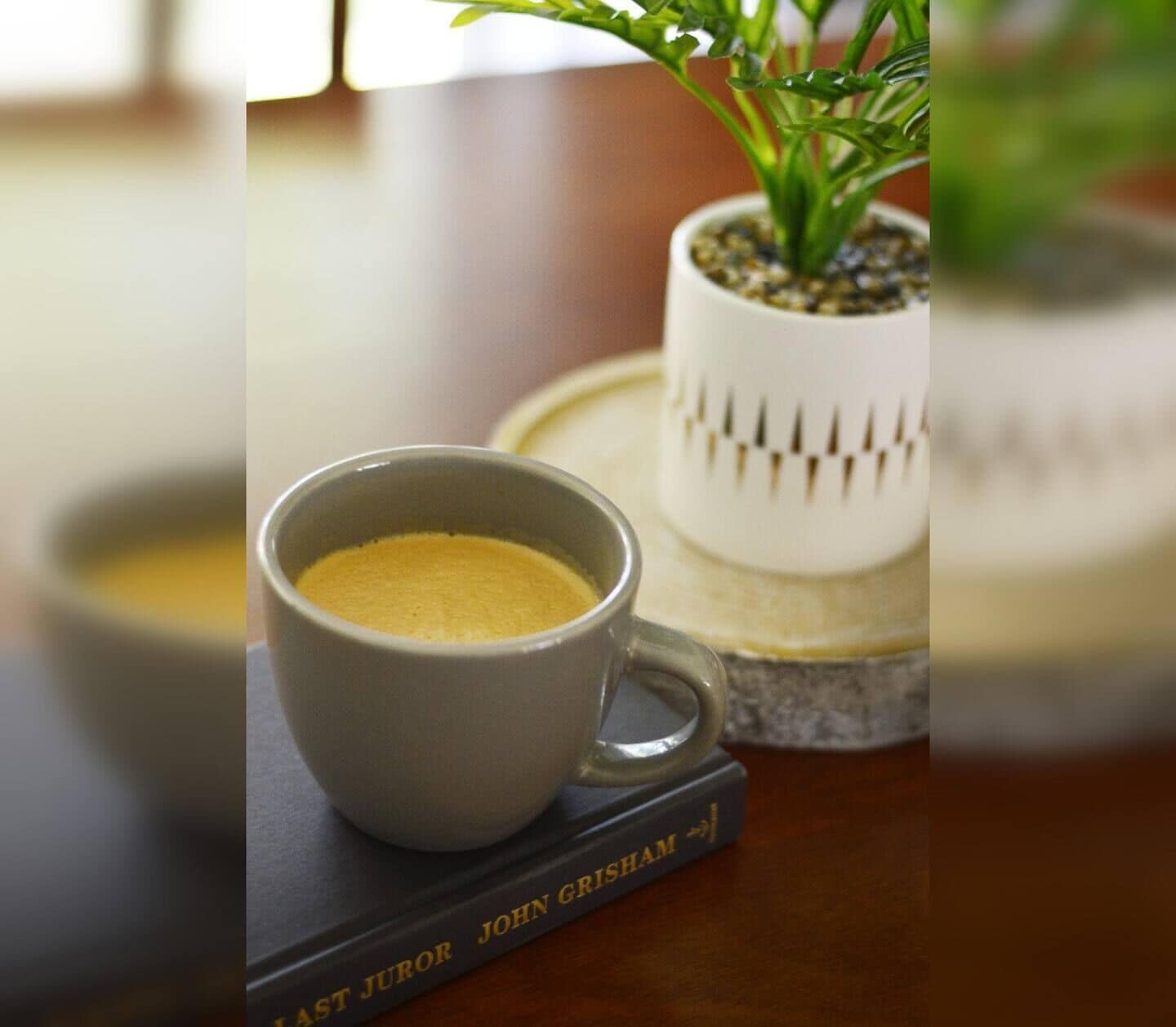A cup of coffee on a table next to a book titled "Last Juror John Grisham" and a small potted plant.