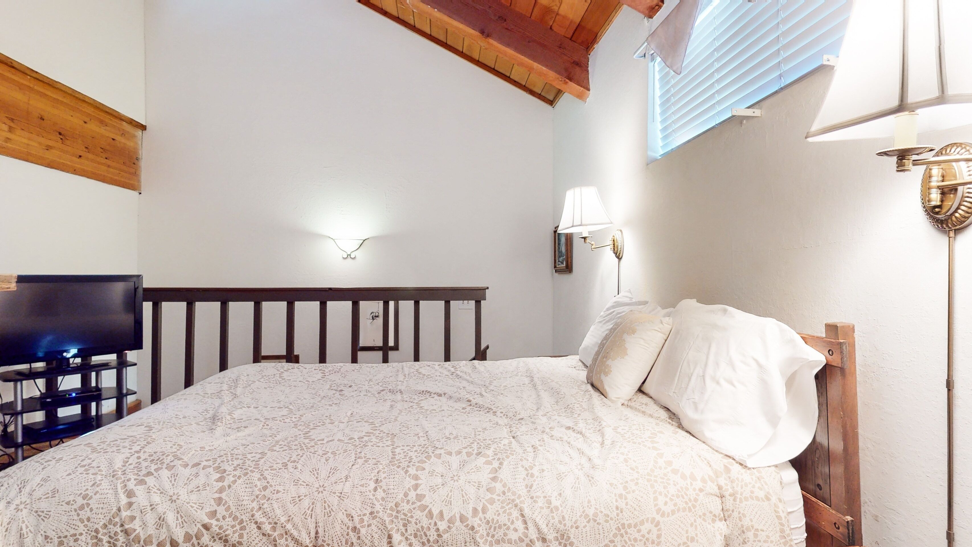 Un dormitorio con cabecera de madera, televisor sobre un mueble bajo, cama con cubrecama de encaje blanco, lámpara de pared y ventana con persianas.