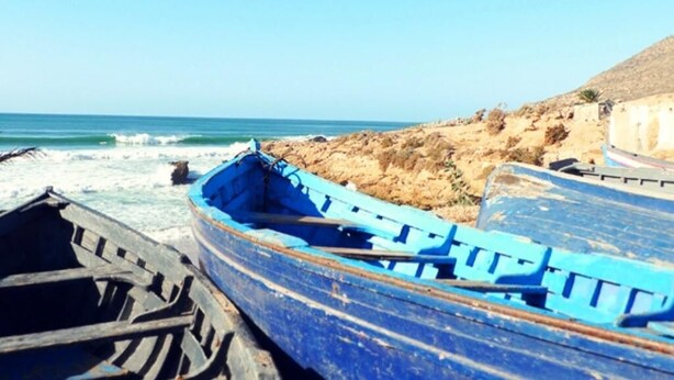 Een strand met twee boten, een blauw en een houten, en een rotsachtige kustlijn.