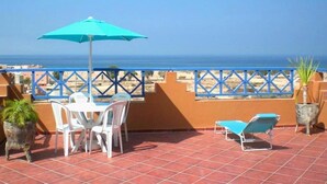 Een terras met een tafel en stoelen, een blauwe parasol en uitzicht op het strand.