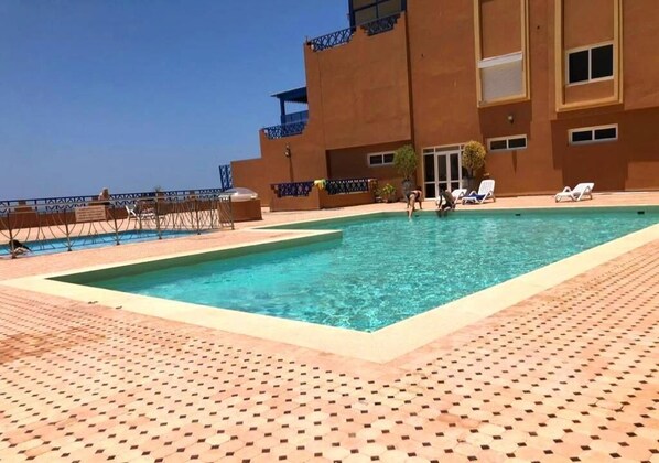Ein Schwimmbecken mit Fliesenbord, ein Gebäude mit Balkonen und Liegestühle.