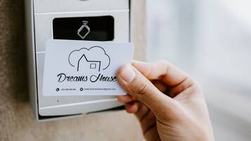 Uma mão segura um cartão com o texto "Dreams House" e um número de contato, em frente a um dispositivo identificado como "safire" com uma câmera e um símbolo de Wi-Fi.