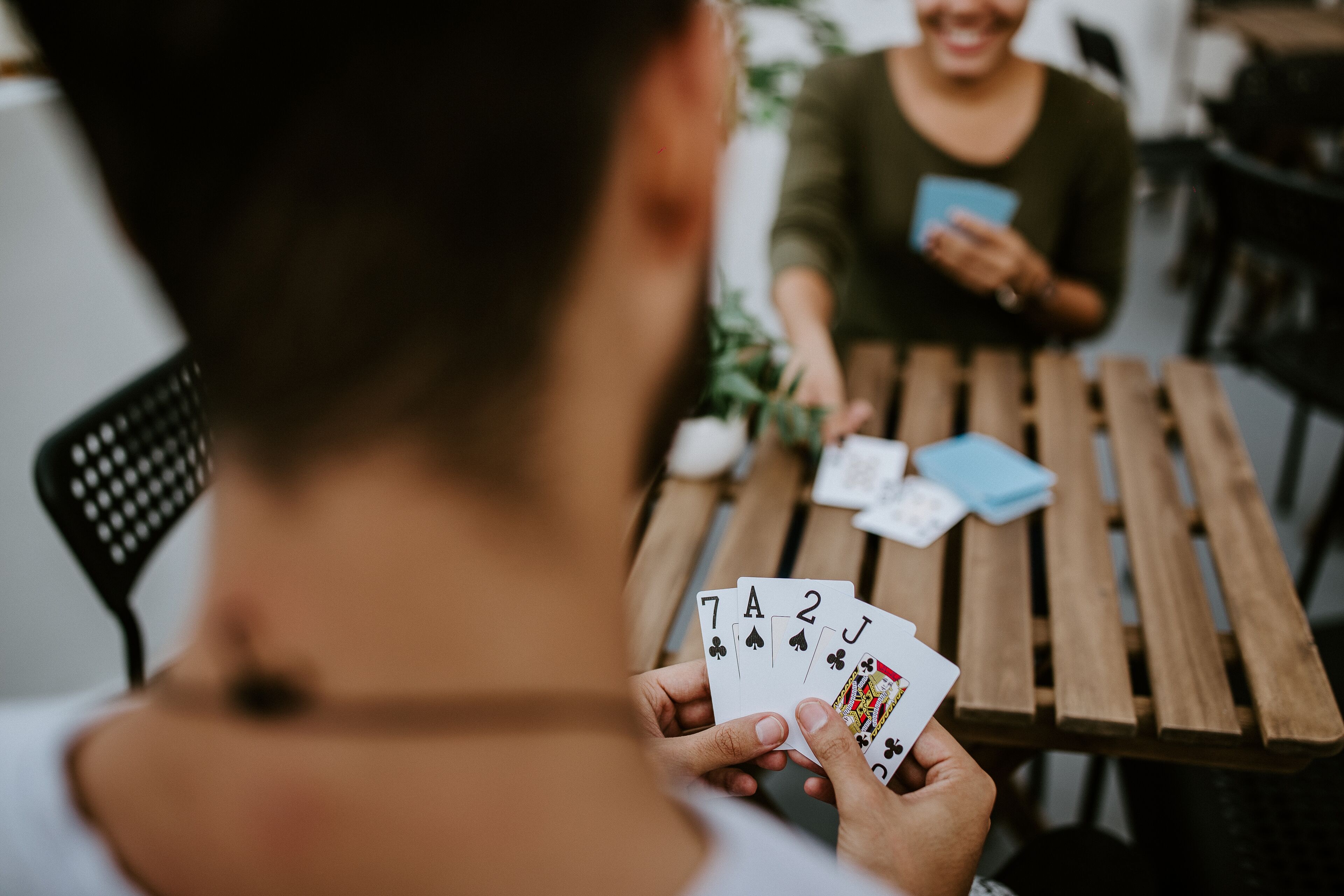 Una persona sosteniendo cartas de juego sobre una mesa de madera, con otra persona sentada al otro lado.