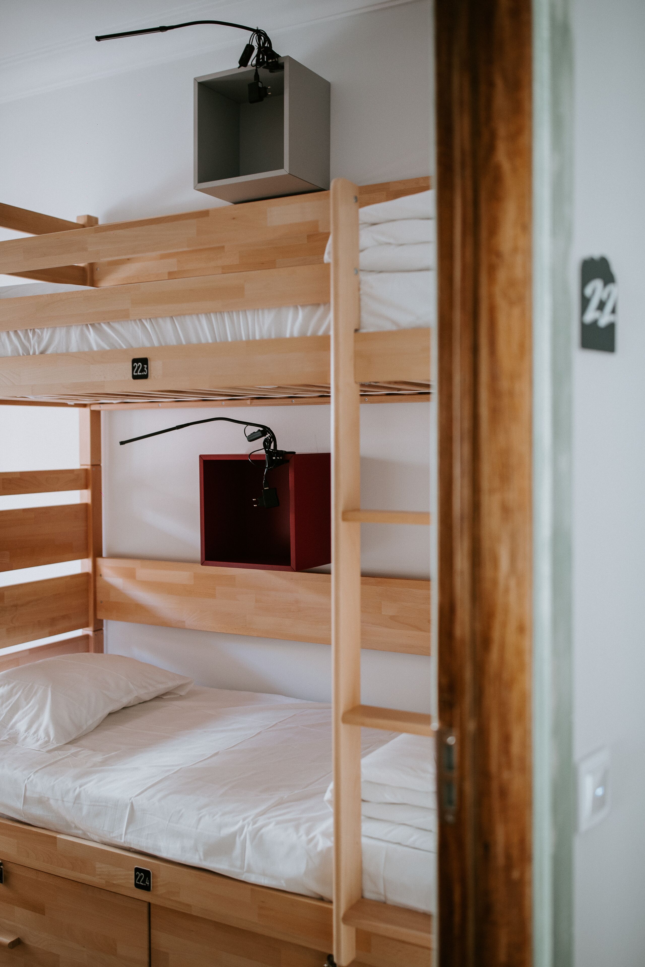 Una cama litera con estructura de madera, un pequeño estante con una caja roja y el número '22' en la pared.