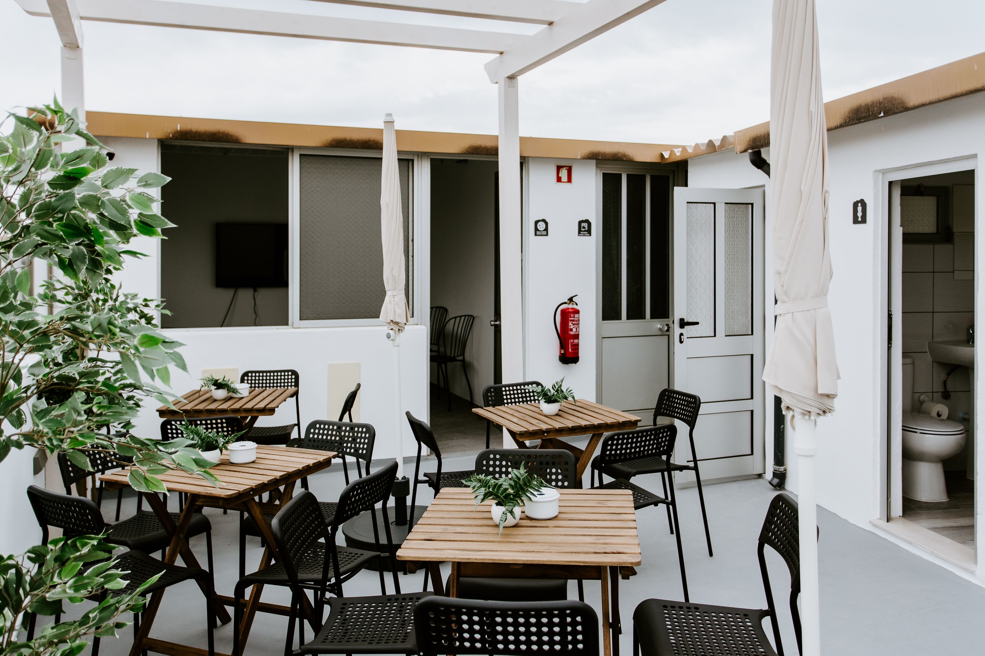 An outdoor patio with wooden tables and black chairs, surrounded by white walls and a fire extinguisher.