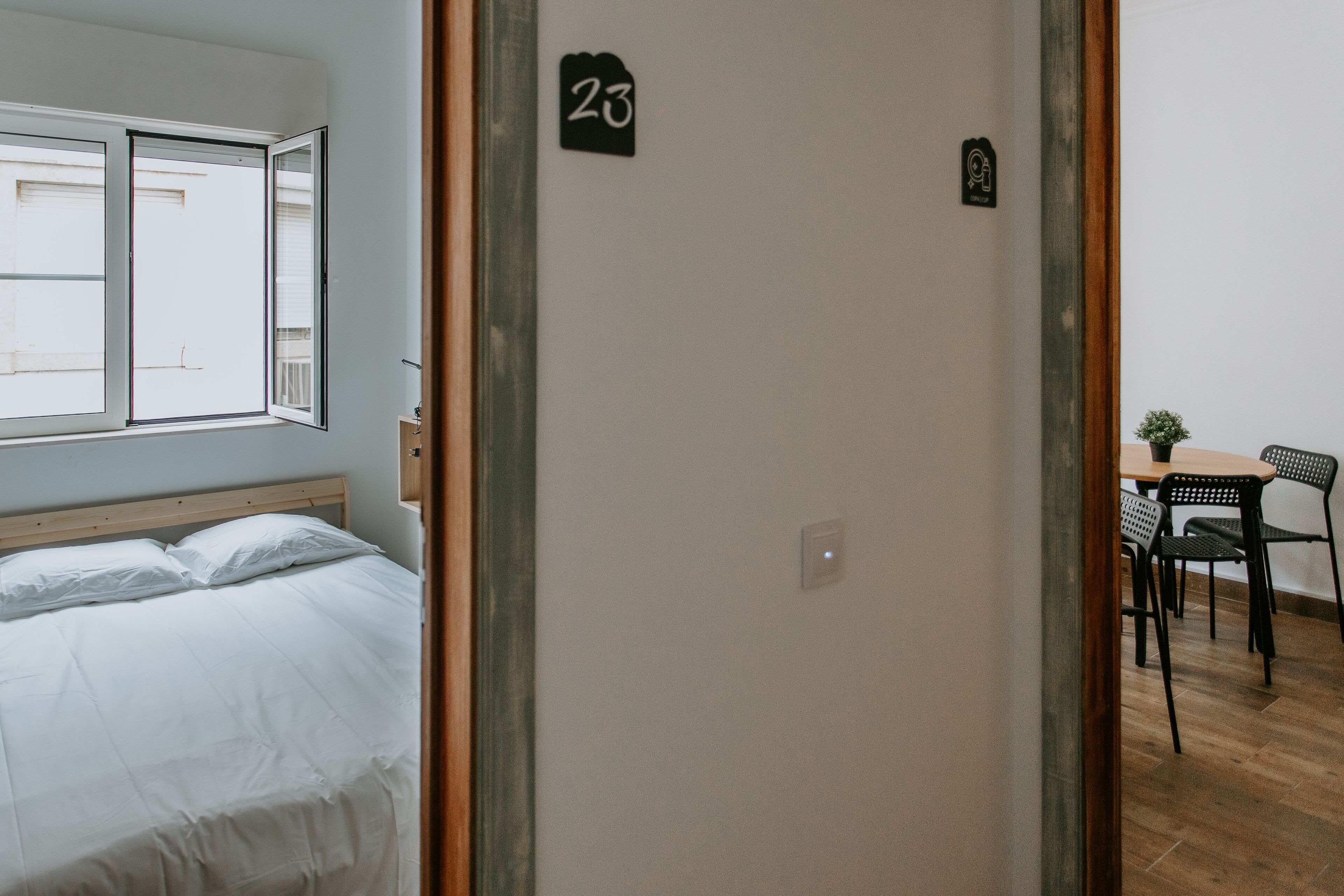Una stanza con un letto, una finestra, una porta con il numero 23 e un tavolo con sedie.