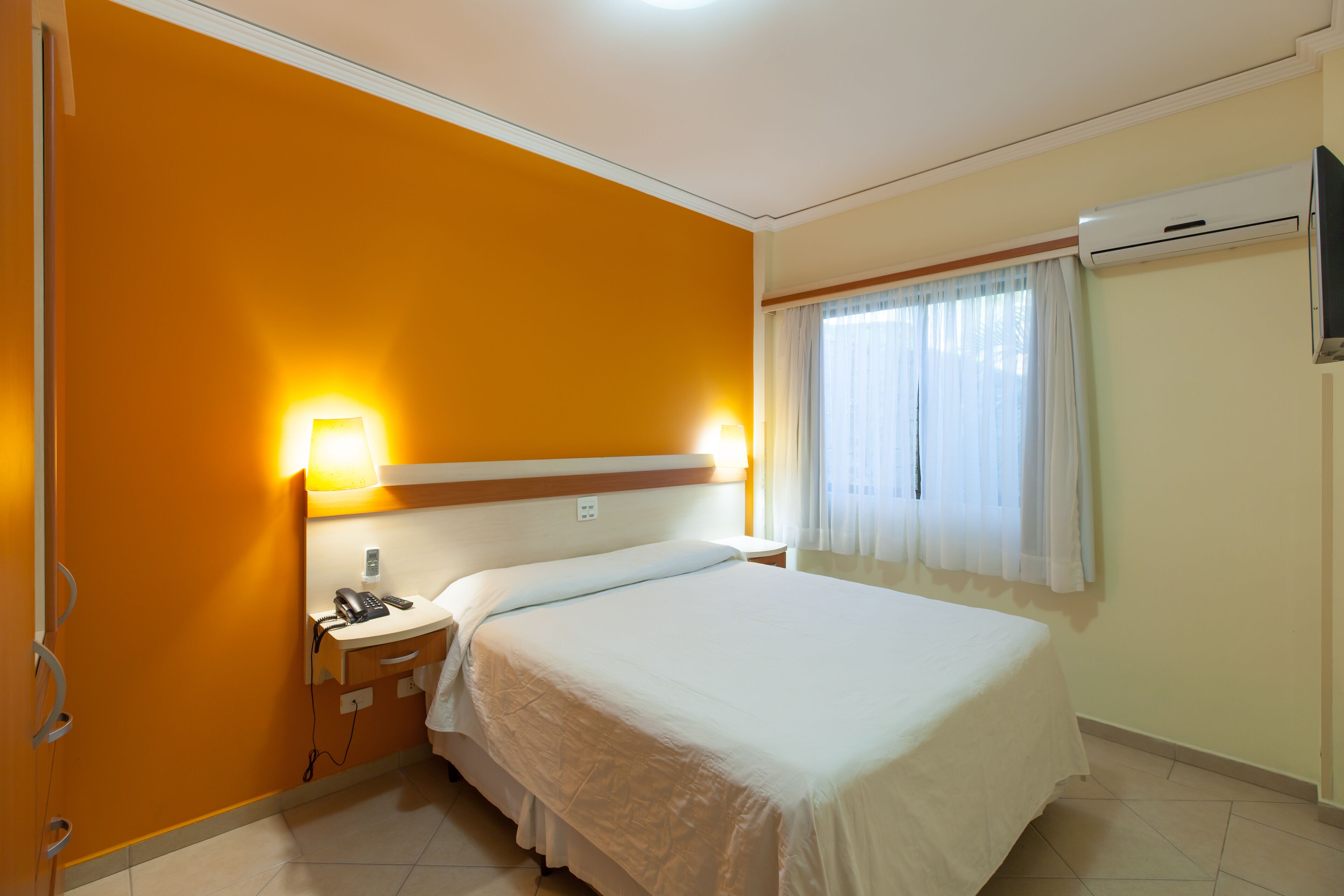 A hotel room with a bed, bedside table, lamp, and an air conditioning unit.
