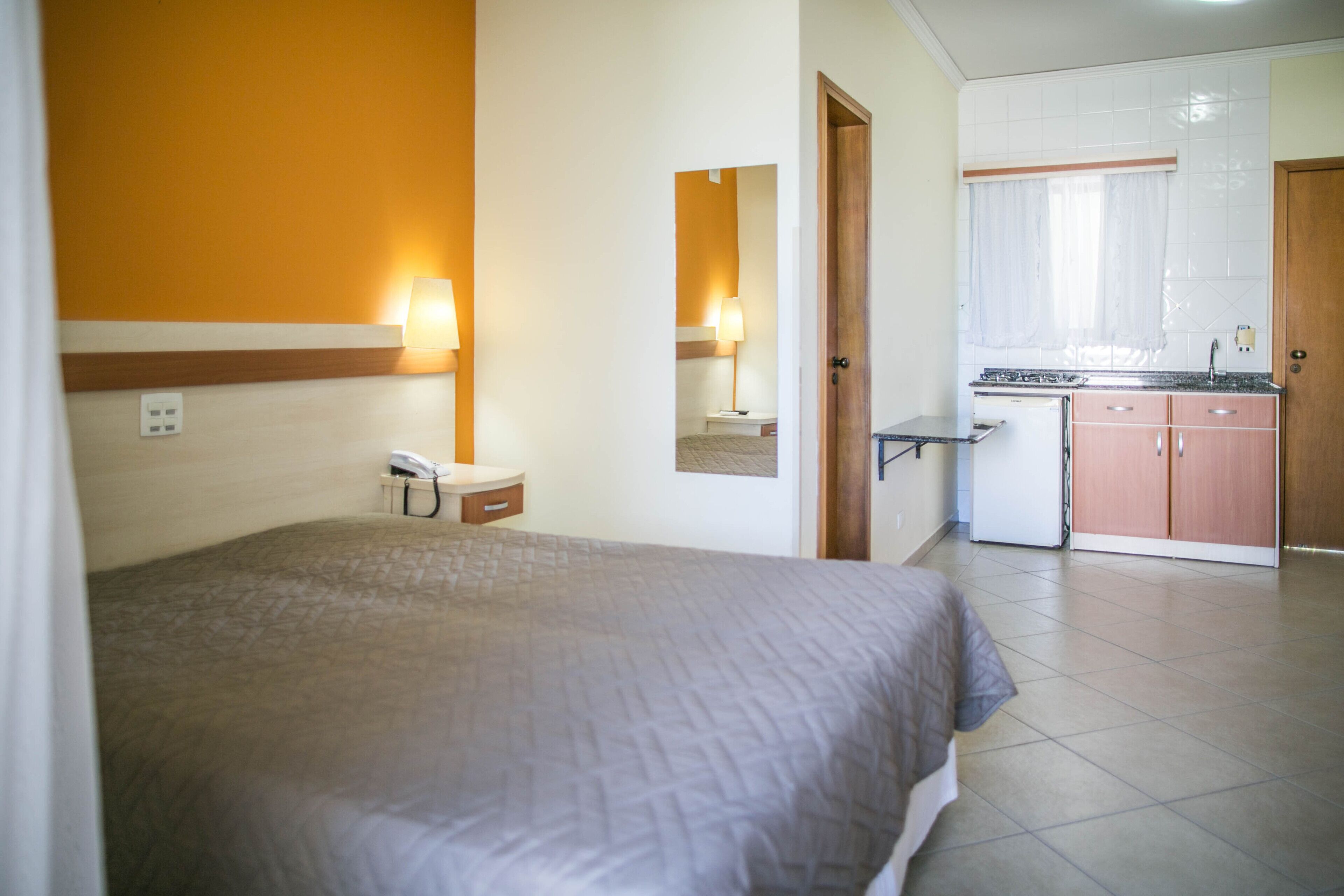 Una habitación de hotel con cama, teléfono en la mesita de noche, un escritorio pequeño con lámpara y una kitchenette con armarios y lavamanos.