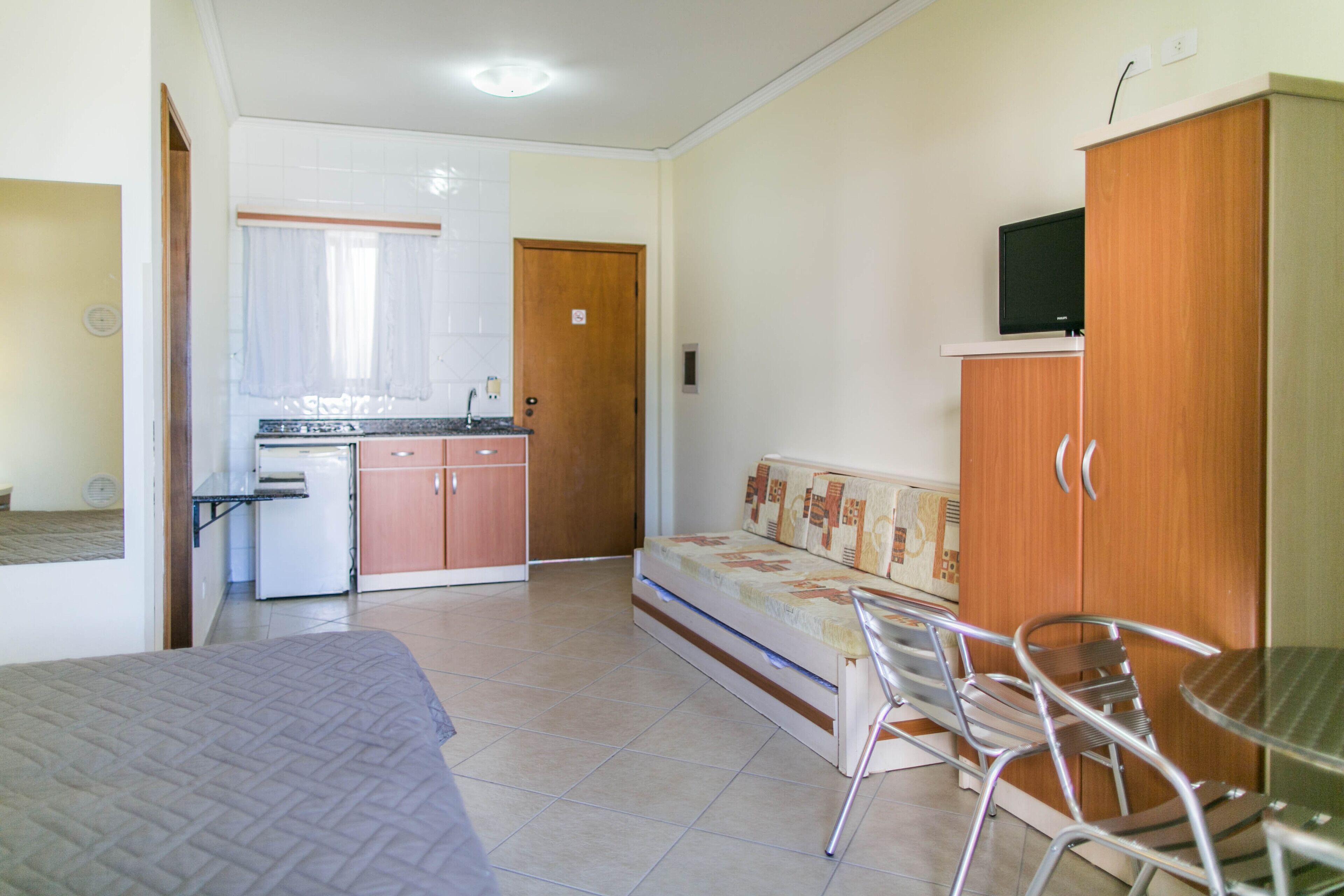 Een kleine kamer met een bed, een slaapbank, een eettafel met stoelen, een kitchenette en een televisie bevestigd aan een kast.