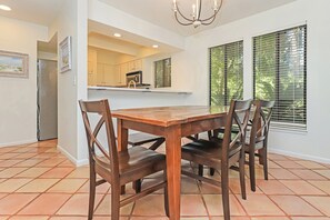 A dining area with a wooden table and chairs, a kitchen in the background, and large windows with blinds.