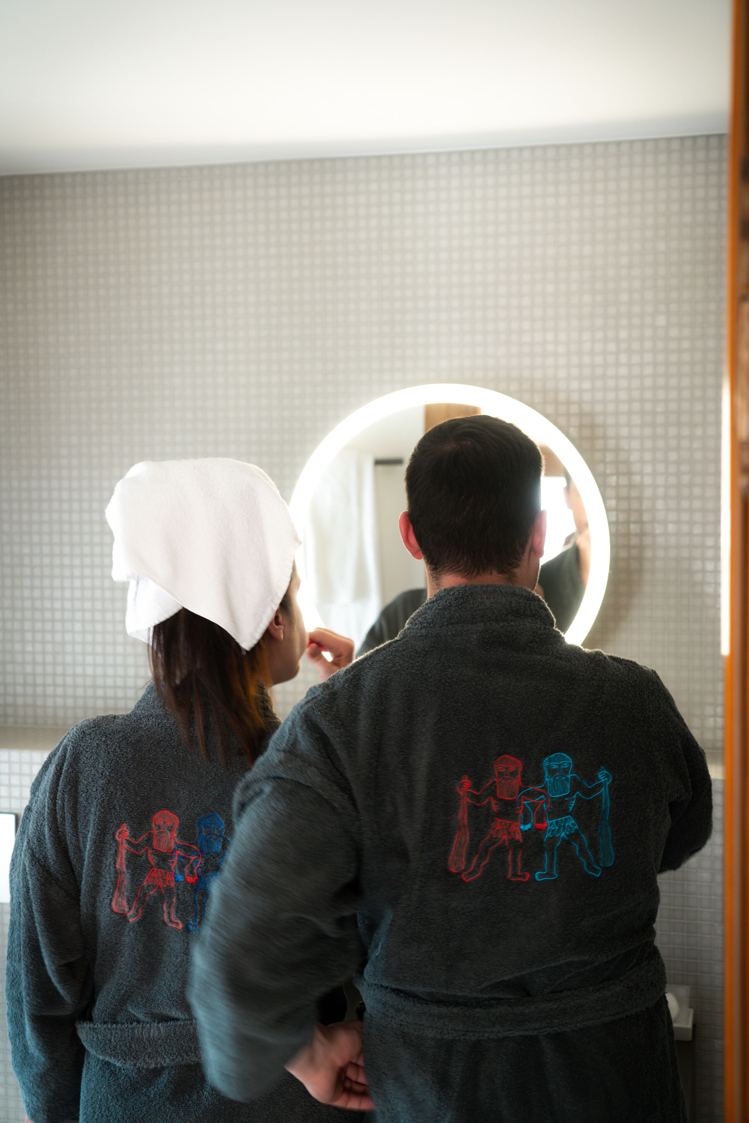 Dos personas en un baño, una con una toalla en la cabeza, ambas vestidas con batas oscuras con un diseño gráfico.