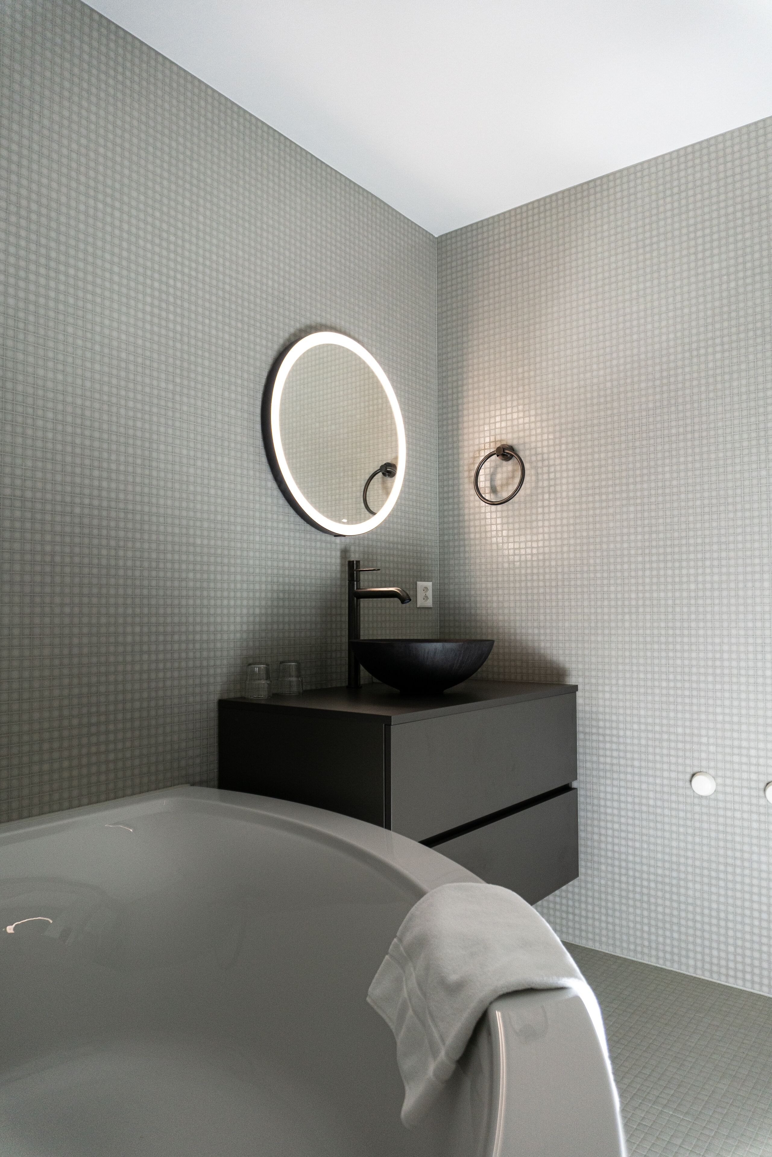 A modern bathroom with a large bathtub, a round mirror with a light, and a black sink.
