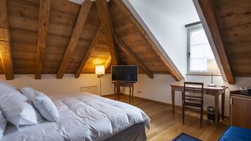 Ein Schlafzimmer mit schrägen Wänden, einem Bett mit weißer Bettwäsche, einem Schreibtisch mit Stuhl und einem Fernseher.