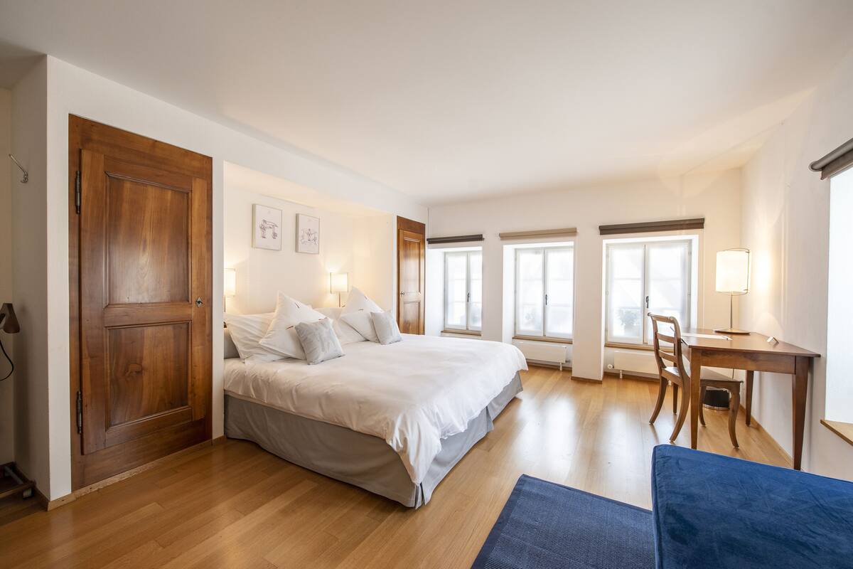 Une chambre avec une porte en bois, un lit avec une literie blanche, un bureau avec une chaise et une fenêtre donnant sur un paysage agréable.