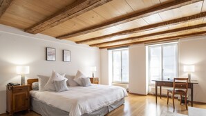 Une chambre avec un plafond en bois, un lit avec une literie blanche, deux lampes de chevet, un bureau avec une chaise et de grandes fenêtres.