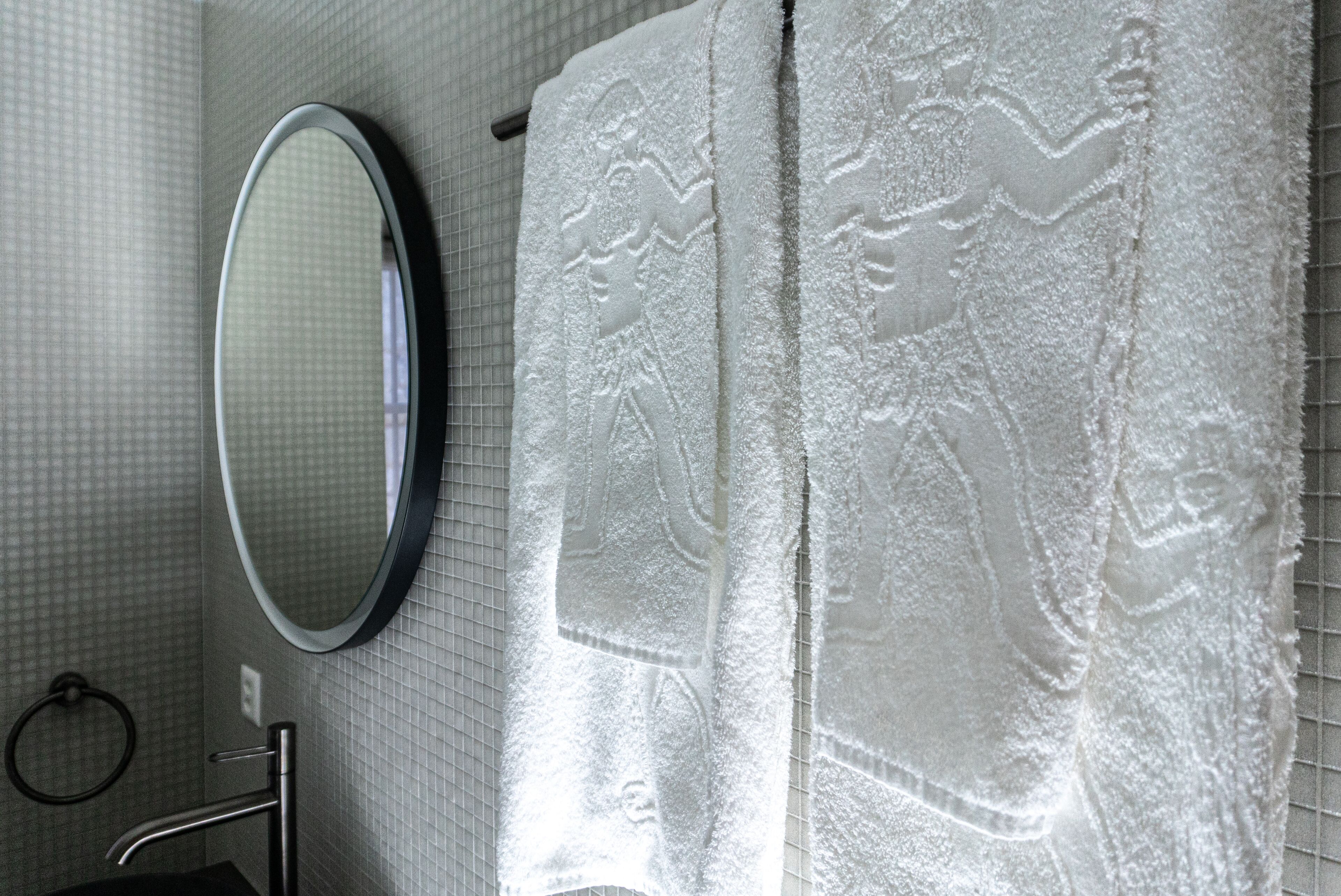 Ein Badezimmer mit gefliesten Wänden, einem runden Spiegel, einem Handtuchhalter mit einem weißen Handtuch und einem chromfarbenen Wasserhahn.