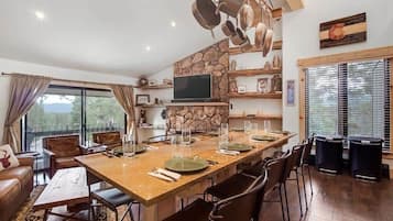 Un vaste espace dédié à la salle à manger, avec une longue table en bois dressée pour quatre personnes, entourée de fauteuils en cuir. On aperçoit un mur en pierre et un téléviseur au fond.