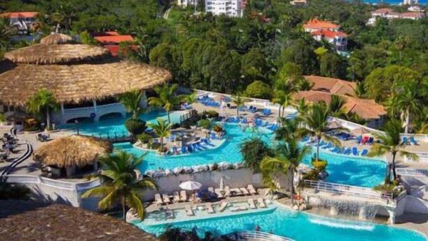 A resort with a large pool area, palm trees, and thatched-roof structures. - Happy As A Clam (Puerto Plata)