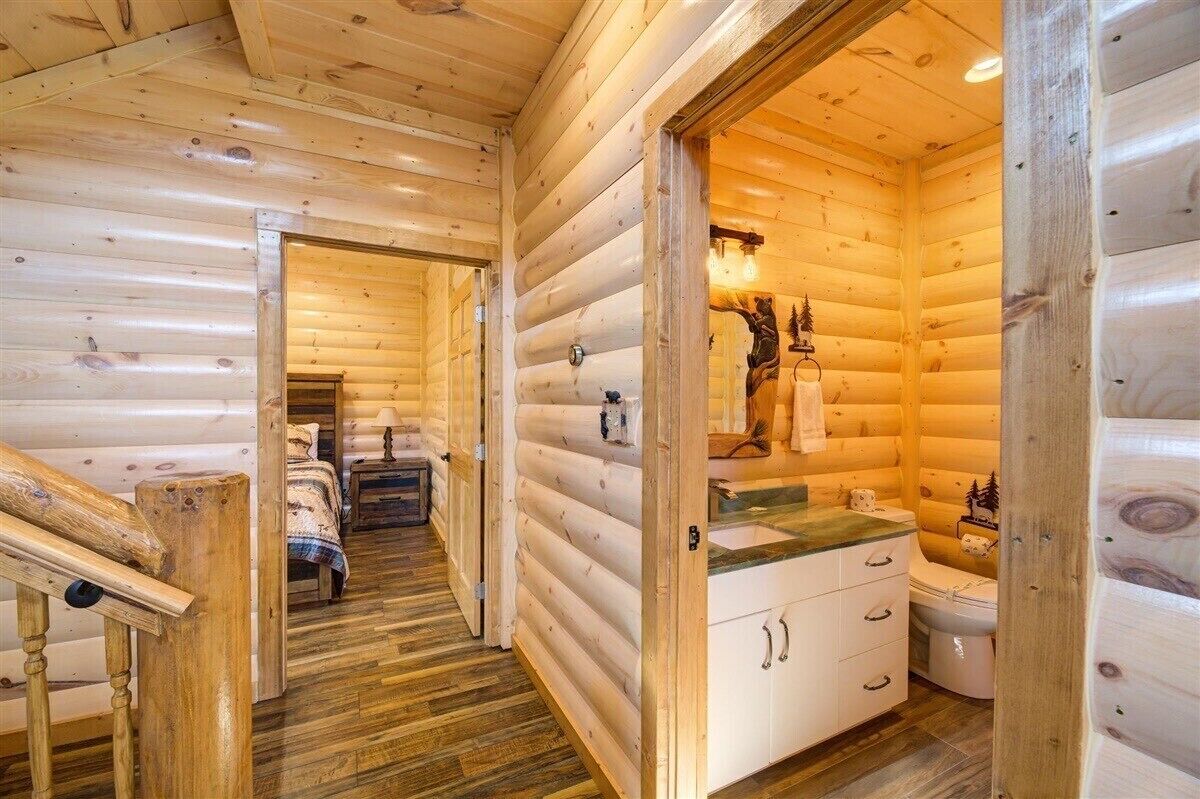 A log cabin interior with a wooden staircase, a bed, a bathroom with a sink and toilet, and a hallway leading to another room.