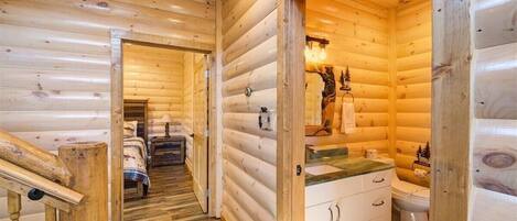 A log cabin interior with a wooden staircase, a bed, a bathroom with a sink and toilet, and a hallway leading to another room.