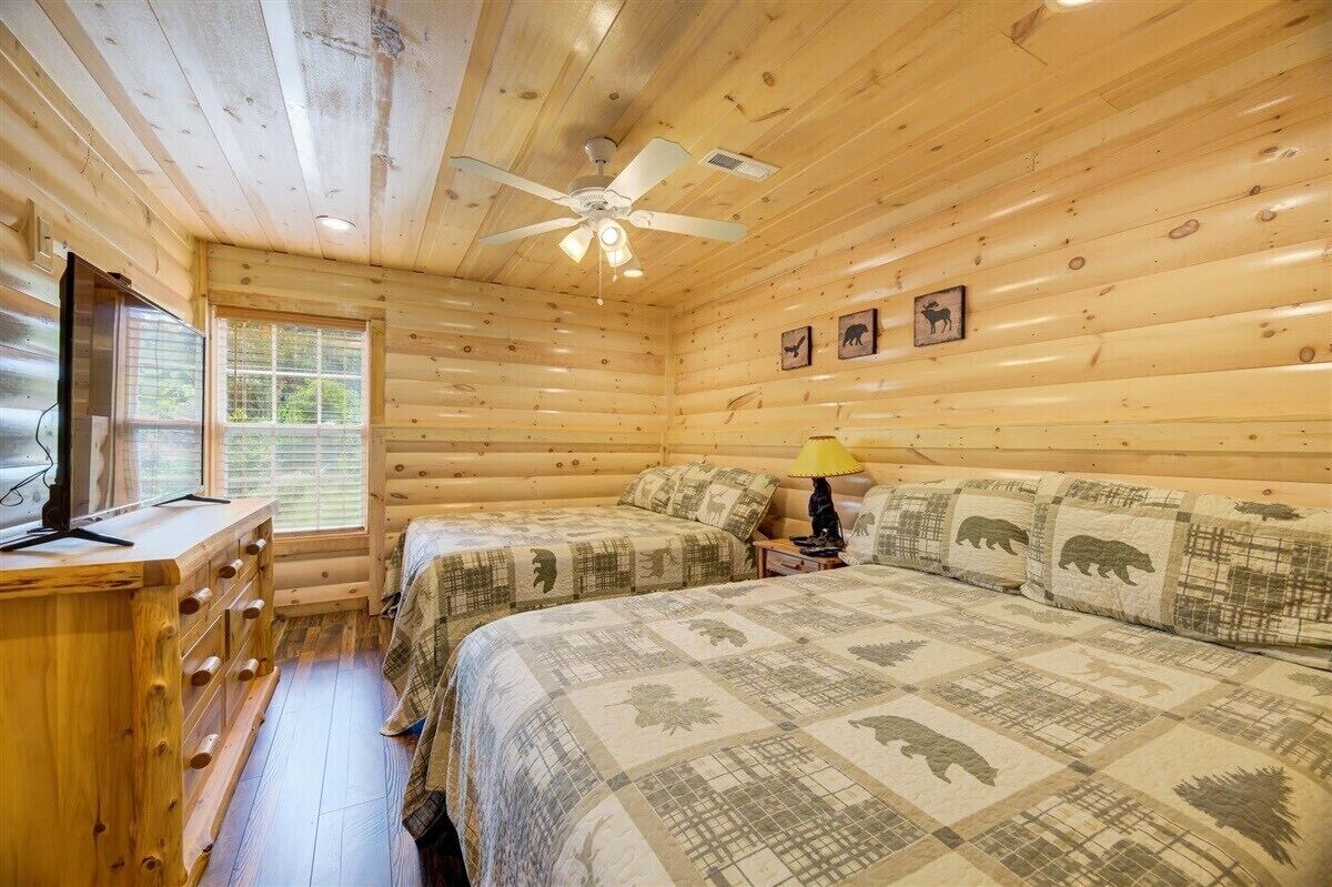 Un dormitorio de cabaña de troncos con dos camas, un ventilador de techo, una ventana y un aparador de madera.