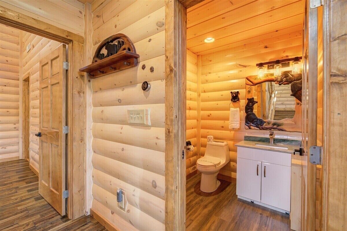 A log cabin interior with a wooden floor, a wooden door, a white toilet, and a small shelf with a mirror and decorative items.