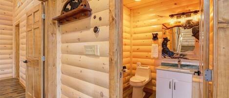A log cabin interior with a wooden floor, a wooden door, a white toilet, and a small shelf with a mirror and decorative items.