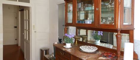 A wooden cabinet with glass doors containing kitchenware, a white vase with purple flowers, and a small trash bin.