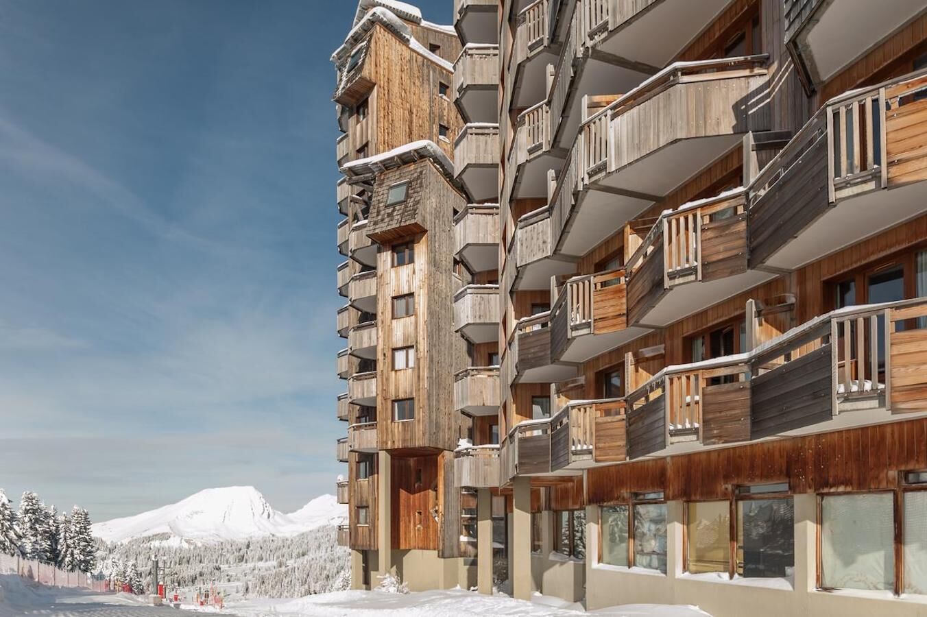 Un hotel de varios pisos con balcones, fachada de madera y un paisaje montañoso nevado al fondo.