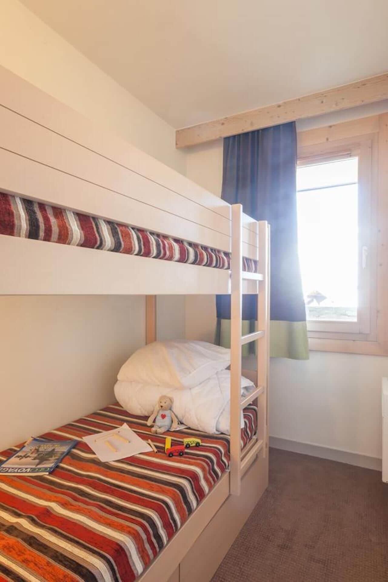 A bunk bed room with striped bedding, a teddy bear, and a window with curtains.