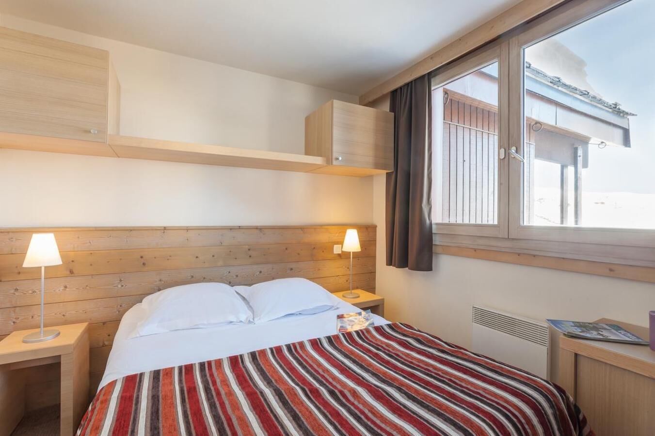 Una habitación de hotel con cabecera de madera, colcha a rayas, dos lámparas de noche, una ventana con cortinas y vista a un edificio.