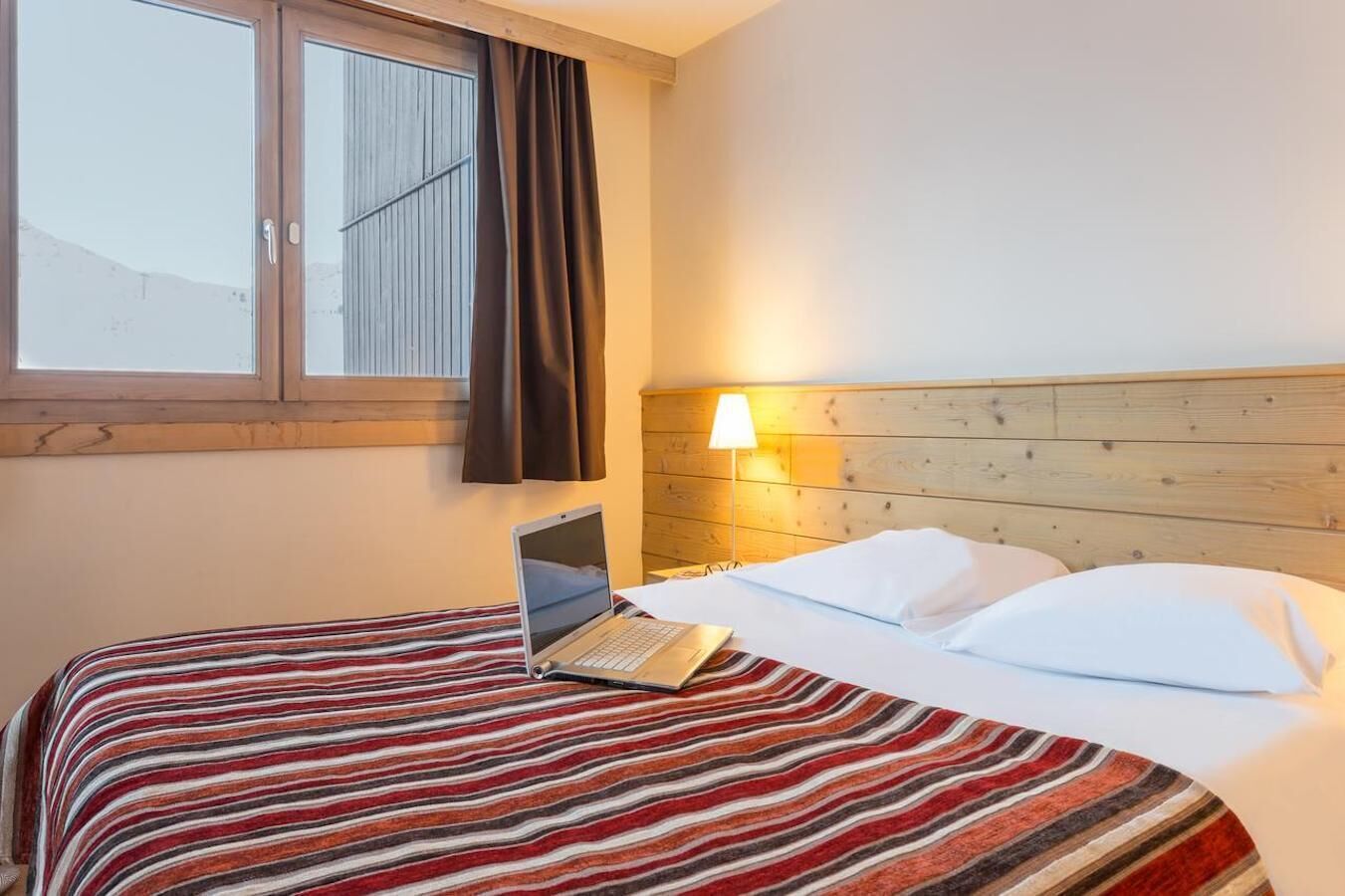Una camera d'hotel con un letto, un portatile sul letto, una finestra con tende e una testiera in legno.