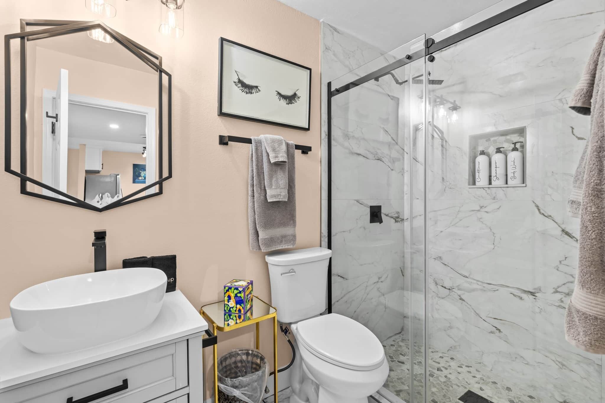 A bathroom with a large oval sink, a toilet, a glass shower enclosure, a framed picture on the wall, and a towel rack.