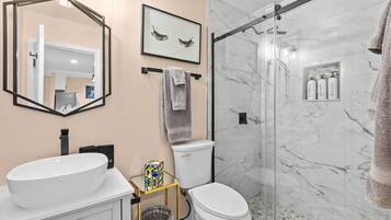 A bathroom with a large oval sink, a toilet, a glass shower enclosure, a framed picture on the wall, and a towel rack.