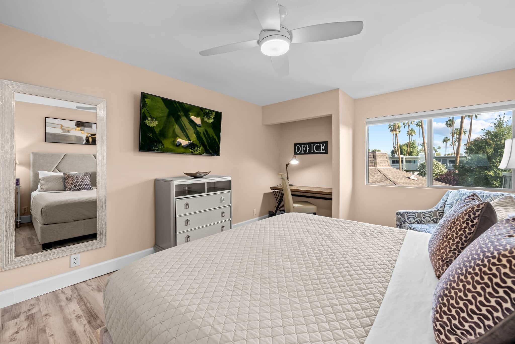 A bedroom with a bed, a desk, a chair, a mirror, and a view of palm trees.