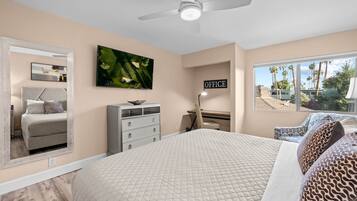 A bedroom with a bed, a desk, a chair, a mirror, and a view of palm trees.