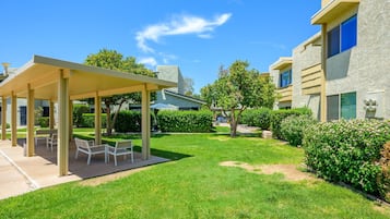 A covered outdoor seating area with tables and chairs, surrounded by well-maintained grass and hedges.