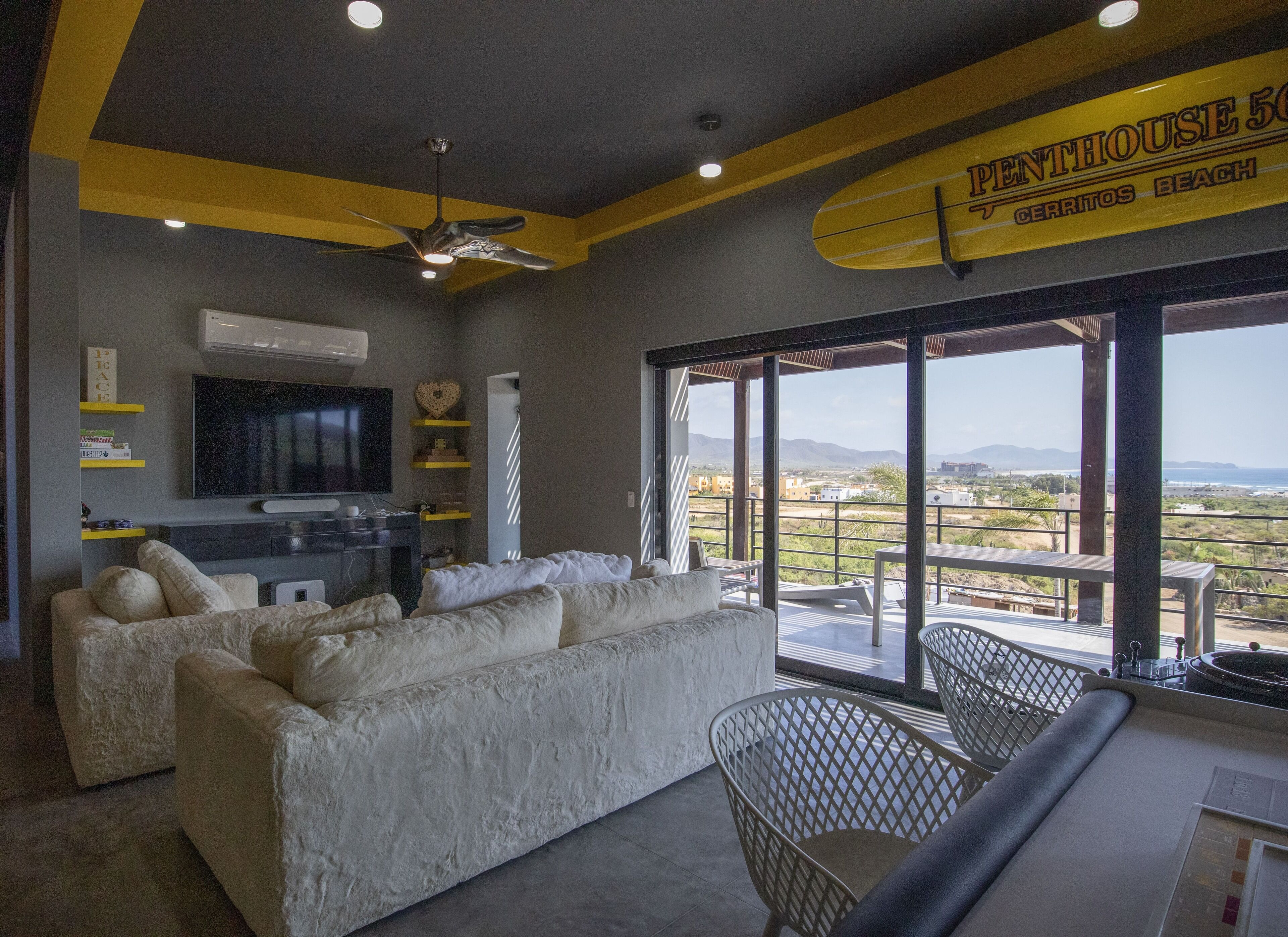 A modern living room with a large sofa, a flat-screen TV, and a ceiling fan. There is a view of the beach and mountains through large windows.